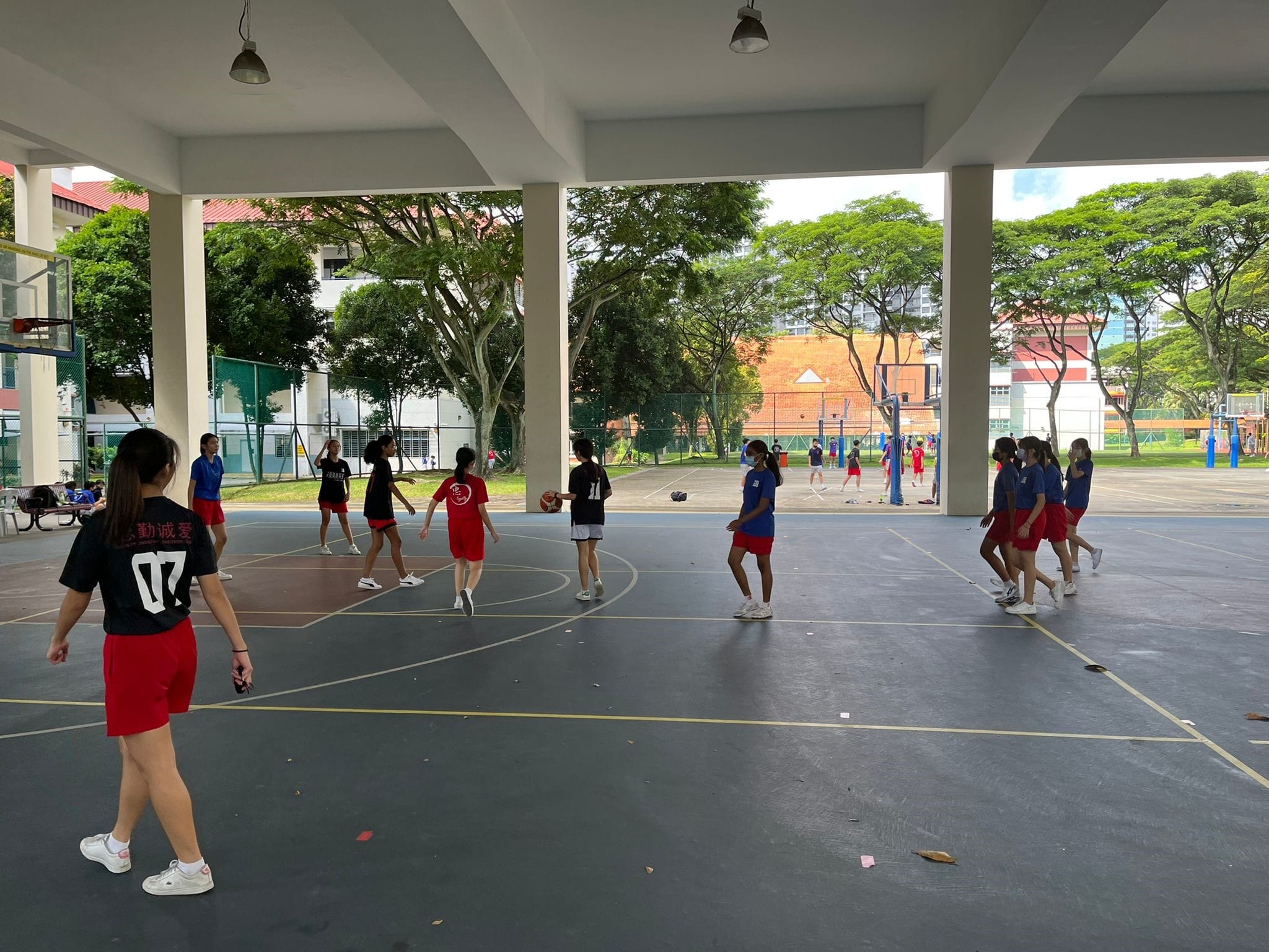Girls in red and blue uniforms play basketball on a covered outdoor court surrounded by trees and buildings.